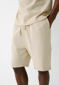 Shorts en coton beige avec une surface texturée, taille élastique, cordon de serrage ajustable et poches latérales, avec une coupe décontractée.