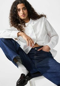 Una camicia bianca con colletto, abbinata a jeans in denim blu scuro. Dettagli includono maniche arrotolate e mocassini neri con una finitura lucida.