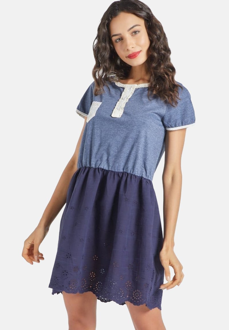 Una mujer sonriente con un vestido azul marino, parte superior de denim, con un borde de encaje y fondo bordado, posando con confianza con los brazos ligeramente levantados.