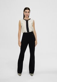 Blusa sin mangas en color crema con un tapón frontal negro, combinada con pantalones de tiro alto negros acampanados y tacones metálicos de punta afilada.