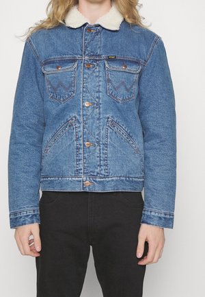 Persona que lleva una chaqueta vaquera azul con cuello de forro polar blanco y botones marrones, combinada con pantalones negros, de pie frente a un fondo liso.