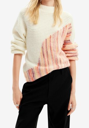 Vrouw draagt een geribbelde crèmekleurige trui met een diagonaal patroon van roze en oranje strepen, gecombineerd met een zwarte broek en zilveren ringen aan haar vingers.