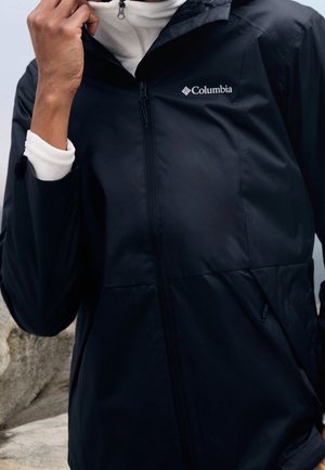 Persoon die een zwarte Columbia-jas draagt over een witte fleece, houdt de kraag van de jas dicht bij de kin, met een rotsachtige achtergrond zichtbaar.