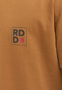 Ruda medvilninė marškinėliai su juodu stačiakampiu logotipu, kuriame užrašyta "RD DO", ir raudonu apskritimu. lygi tekstūra, trumpomis rankovėmis, apvaliu kaklu.