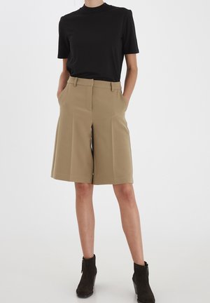 Persoon met beige, op maat gemaakte shorts tot op de knie, een zwart T-shirt met korte mouwen en zwarte enkellaarsjes, staand met handen in de zakken.