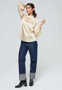 Blouse en satin crème avec un col haut et un petit détail en forme de trou, associée à un jean en denim bleu foncé avec des ourlets retroussés et des bottes à talons carrés noires.