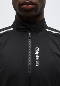 Primer plano de una persona que lleva una chaqueta negra con cremallera y el logo "GripGrab" en blanco en el pecho, con franjas reflectantes en los hombros.