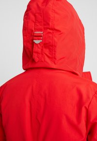 Rood waterdicht jack met capuchon, voorzien van een aansnoerband en metalen accenten. De stof heeft een gladde, licht glanzende afwerking.