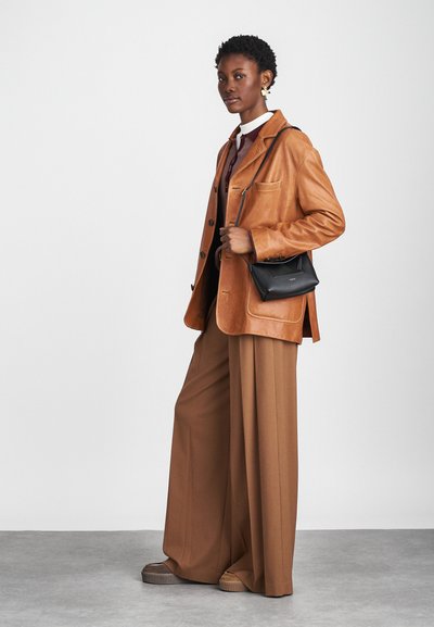 Chaqueta de cuero marrón con bolsillos delanteros, sobre un suéter burdeos y una camisa blanca. Pantalones beige sueltos y un bolso cruzado negro completan el look.