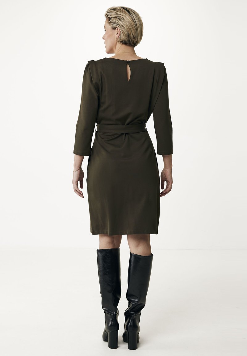 Oliwkowa sukienka do kolan z długimi rękawami i wycięciem w kształcie klucza na plecach. Połączona z wysokimi, błyszczącymi, czarnymi kozakami do kolan.