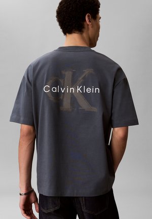 Mand iført mørkegrå T-shirt med stort falmet "CK" og hvid "Calvin Klein" tekst på ryggen, stående mod en ensfarvet grå baggrund.
