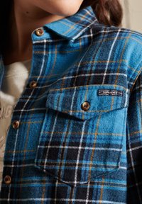 Camicia flanella a quadri in toni di blu, nero e giallo, con chiusura a bottoni frontale, taschino sul petto e etichetta con logo sulla tasca.