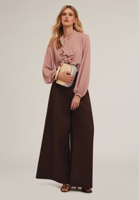 Ljust rosa blus med volanger och långa ärmar, matchad med mörkbruna vida byxor. Modellen håller en liten tvåfärgad handväska.