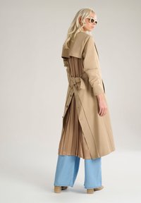 Trench coat beige con un retro marrone plissettato, legato in vita con un fiocco. Il modello indossa pantaloni azzurri a gamba larga e scarpe beige.