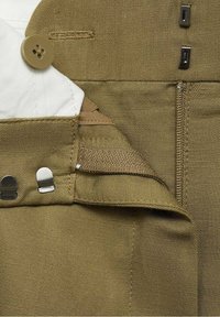 Gros plan sur la braguette d'un pantalon kaki avec bouton, fermeture à crochet et barre, et coutures visibles sur le tissu.