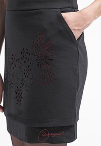 Schwarzer Baumwollrock mit floralen Cut-out-Mustern in Rot, mit Bund und Seitentasche. Der Saum ist leicht geschwungen.