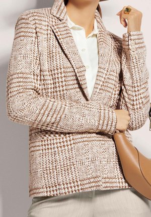 Kobieta ubrana w brązowo-biały blezer w kratę pstrokatą na białej koszuli, trzymająca brązową skórzaną torebkę z dużym pierścieniem na palcu.