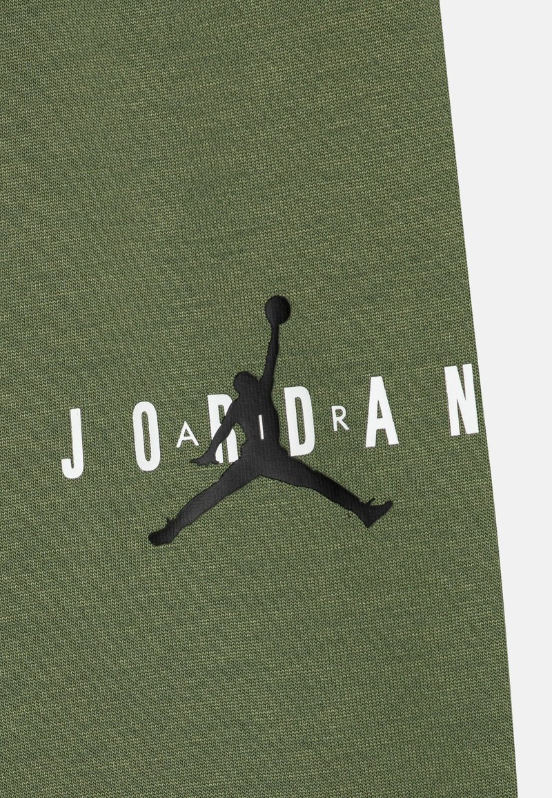 Silhouette nera di Jumpman con pallone da basket sopra il testo "JORDAN" bianco su uno sfondo di tessuto testurizzato verde oliva.