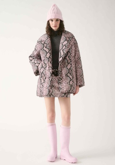 Oversized Jacke mit pinkem Schlangenhautmuster und taillierter Silhouette, getragen über einem grauen Rollkragenpullover und einem passenden Minirock. Pinke Stiefel aus Gummi bis zum Knie.