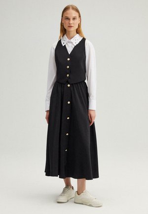 Giovane donna con camicia bianca a maniche lunghe con colletto traforato, gilet nero con bottoni e gonna nera abbinata, sneakers bianche.