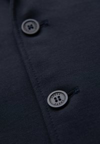 Tissu bleu foncé avec deux boutons ronds cousus, un bouton en gros plan avec le texte « OMBRE » sur son bord.