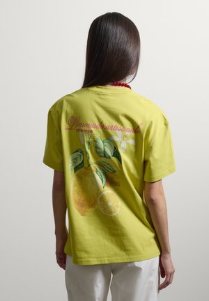 Person mit langen dunklen Haaren, die ein gelbes T-Shirt mit Zitronen und französischem Text trägt, kombiniert mit weißen Hosen, vom Kamerablick abgewandt.