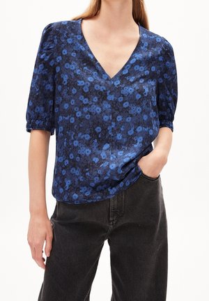 Vrouw die een blauw bloemenblouse met V-hals draagt, voorzien van opgeblazen korte mouwen en zwarte, hooggetailleerde jeans, met haar hand in de zak.