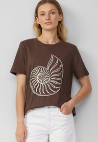 Brązowy bawełniany t-shirt z dużą białą i kolorową grafiką muszli nautilusa. Krótkie rękawy i luźny krój. Sparowany z białymi spodniami.