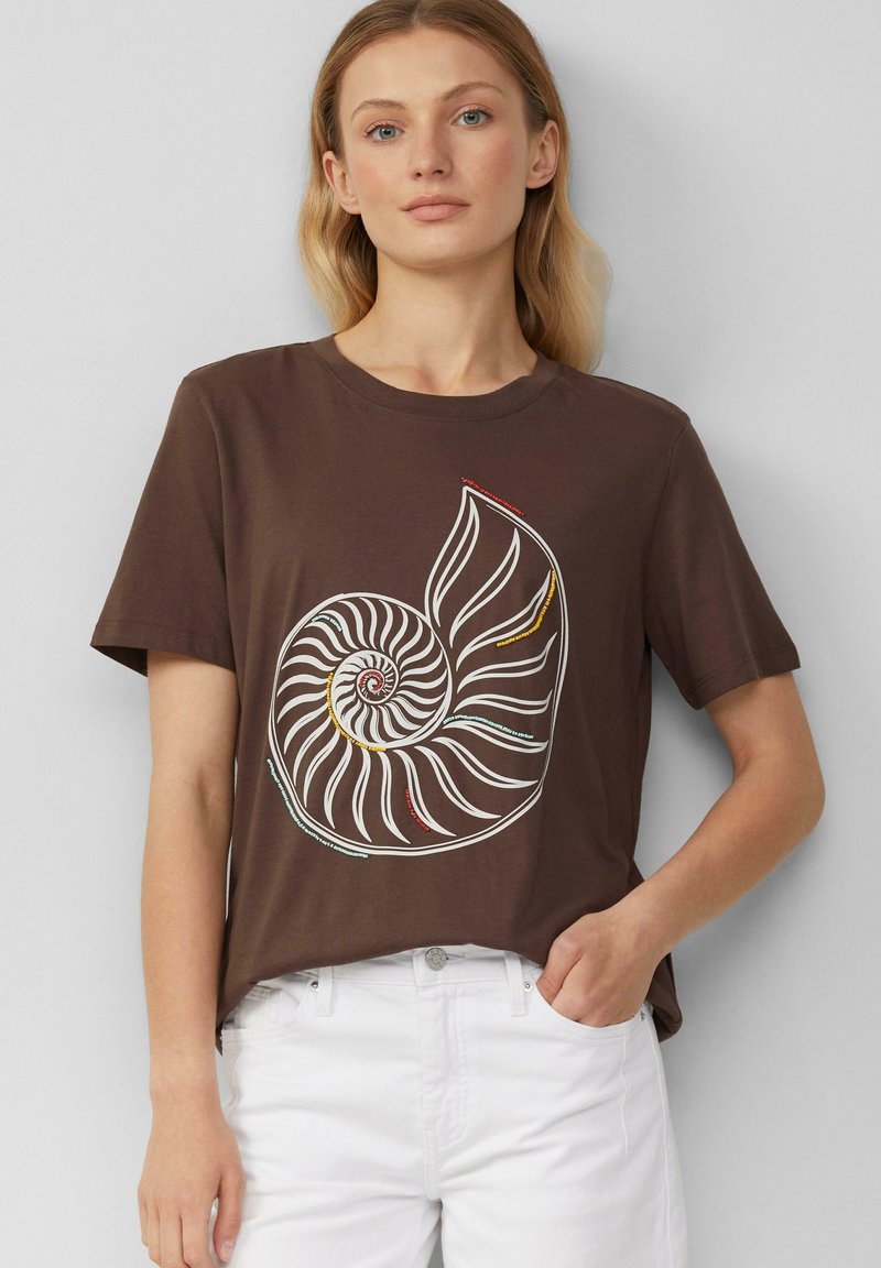 Brązowy bawełniany t-shirt z dużą białą i kolorową grafiką muszli nautilusa. Krótkie rękawy i luźny krój. Sparowany z białymi spodniami.