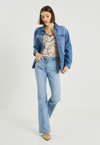 Chaqueta vaquera azul claro, de corte relajado y cierre de botones. Usada sobre una blusa beige con estampado y combinada con vaqueros acampanados.