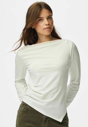 Ung kvinde med langt brunt hår iført en off-white langærmet top og mørkegrønne bukser mod en simpel lys baggrund.