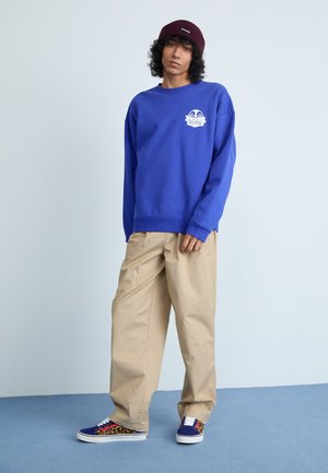 Felpa blu con logo, pantaloni beige larghi e sneakers con stampa leopardata multicolore, completati da un berretto bordeaux.