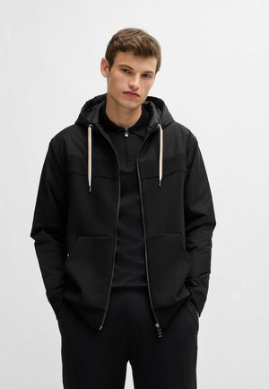Zwarte lichte hooded jack met een ritssluiting aan de voorkant, voorzakken en contrasterende textuurpanelen. Voorzien van koorden met metalen punten.