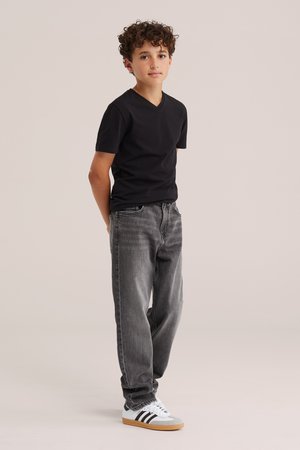 Garçon aux cheveux bouclés portant un t-shirt noir, un jean gris et des baskets blanches, debout les mains dans le dos sur un fond uni.