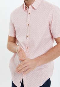Mann mit einem rosa Kurzarm-Hemd mit weißem Blattmuster, der die Hände vor einem weißen Hintergrund faltet.