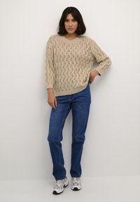 Maglione beige lavorato a maglia con un motivo a onda, taglio morbido e maniche a tre quarti abbinato a jeans blu e scarpe da corsa grigie.