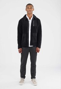 Sort fleecejakke med frontlomme, båret over en hvid skjorte. Modellen har også mørkegrå bukser og lyse sneakers på, mens han står mod en ensfarvet baggrund.