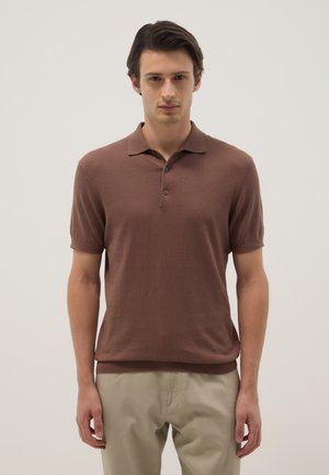 Hombre de pie con una camisa polo marrón de manga corta y pantalones beige sobre un fondo claro y liso.