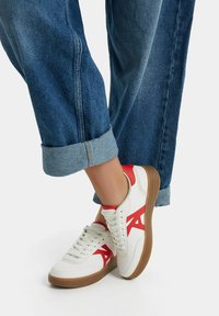 Vita sneakers med röda accenter, rund tå, texturerad gummisula och en design med en stor röd logotyp. Bärs med uppkavlade blå jeans.