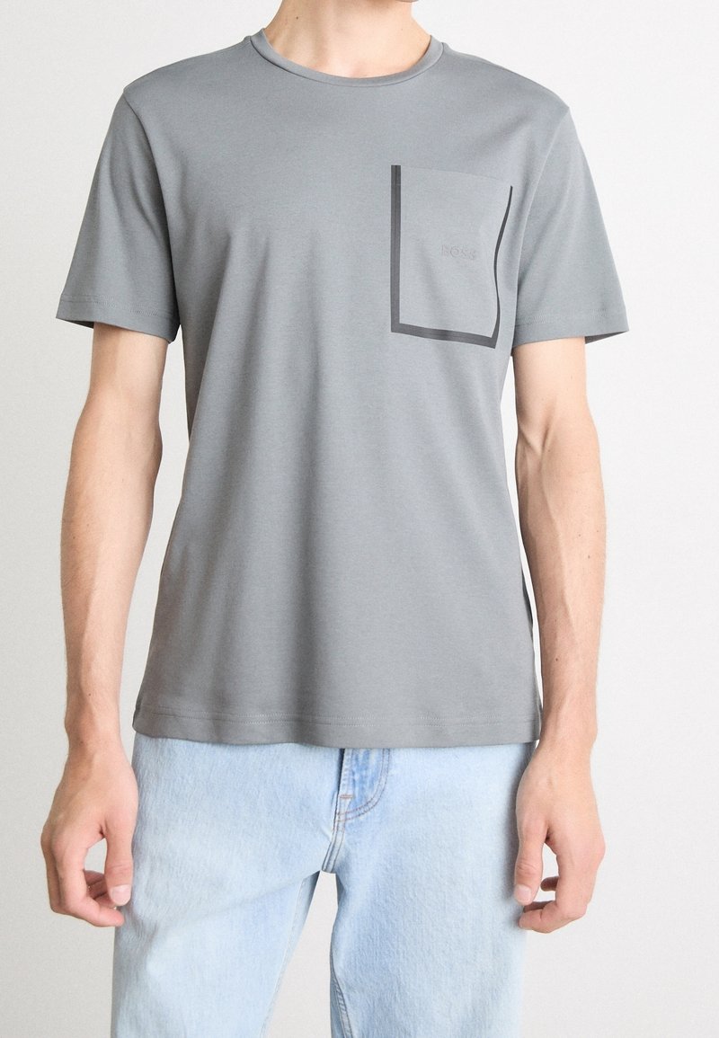 Homme portant un t-shirt gris à manches courtes avec une poche à contour sombre sur le côté gauche de la poitrine et un jean bleu clair.