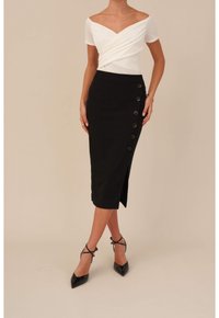 Weißes schulterfreies Oberteil mit verdrehtem Stoffdetail, kombiniert mit einem schwarzen Bleistiftrock mit einem seitlichen Schlitz und sieben Knöpfen zum Schließen. Schwarze spitze High Heels.