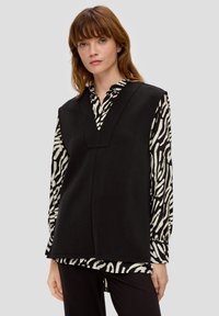 Top nero senza maniche con scollatura a V sopra una camicia a maniche lunghe con stampa zebratica bianca e nera. Tessuto morbido e design comodo.