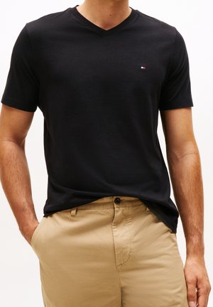 Homme portant un t-shirt noir à manches courtes et col en V avec un petit logo sur la poitrine et un pantalon beige, une main dans la poche.