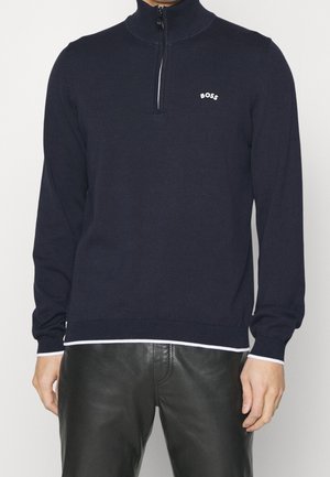 Mannequin portant un pull BOSS bleu marine foncé à fermeture éclair courte, avec une bordure blanche, et un pantalon en cuir noir.