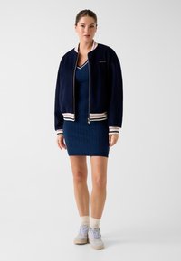 Veste bomber marine avec poignets et ourlet côtelés, ornée d'un logo doré et de détails rayés, associée à une robe tricotée bleue et des baskets claires.