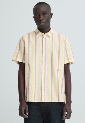 Jeune homme portant une chemise boutonnée à manches courtes avec des rayures verticales beige, jaune et blanches, debout devant un fond uni.