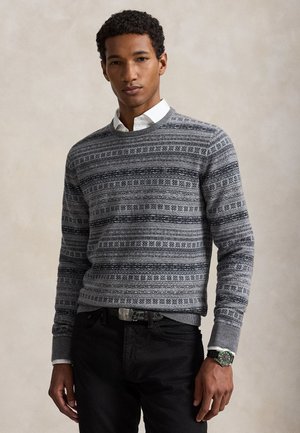 Jeune homme portant un pull gris à motifs par-dessus une chemise blanche avec un pantalon noir et une montre, debout devant un fond beige uni.
