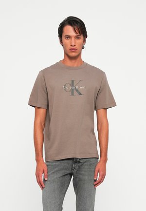 Tânăr îmbrăcat cu un tricou taupe cu logo-ul Calvin Klein și blugi gri, stând în fața unui fundal deschis.