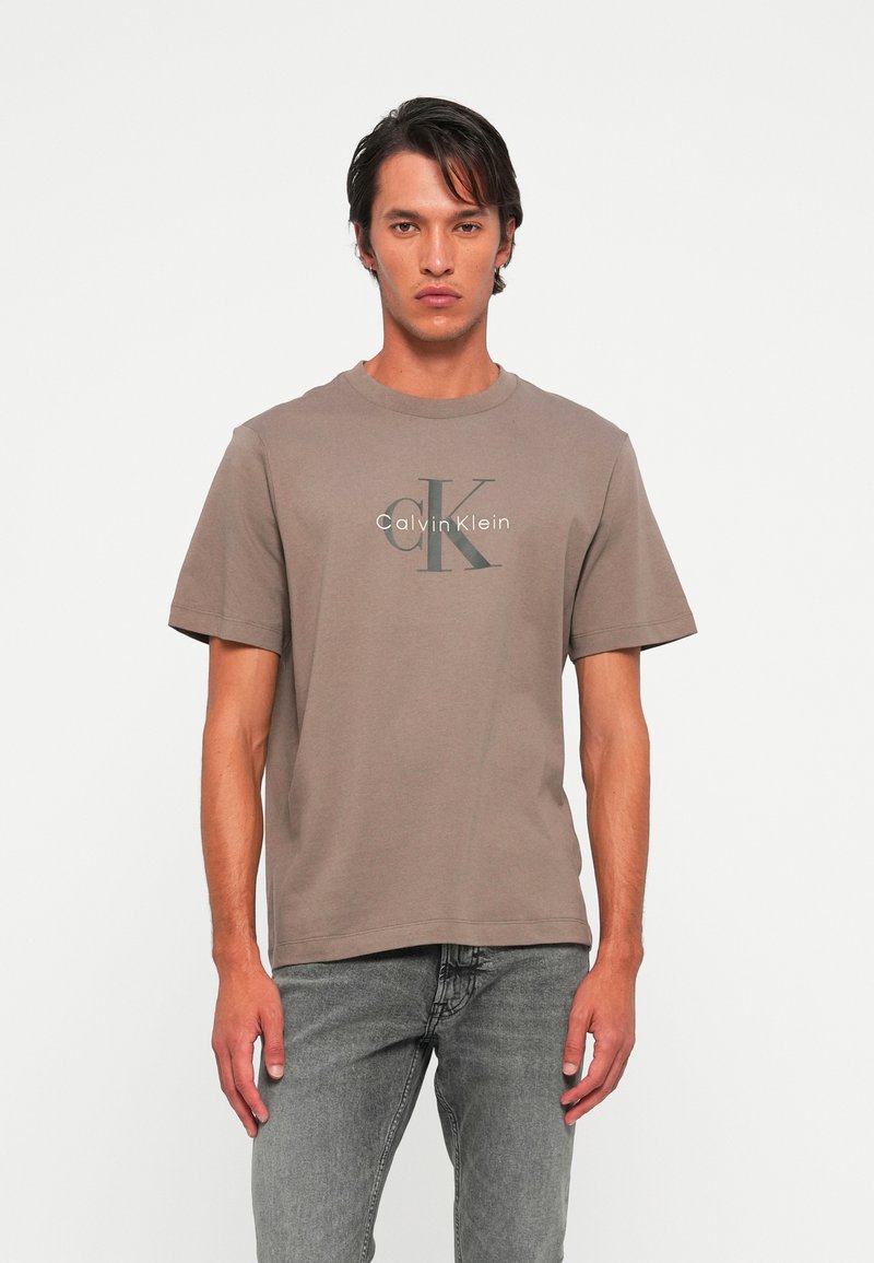 Mladic odjeven u taupe majicu s logotipom Calvin Klein i sive traperice, stoji naslonjen na jednostavnu svijetlu pozadinu.