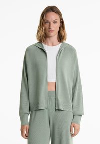 Mintgrön zip-up kofta med ribbad textur, huva och sänkta axlar. Bärs över en vit cropped top, i kombination med matchande byxor.